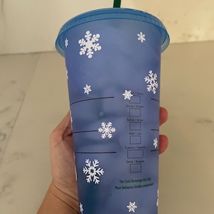 Puede incluir: Un vaso azul reutilizable para bebidas fr&iacute;as con dise&ntilde;os de copos de nieve blancos. El vaso tiene una pajita verde y una tapa azul. El texto en el vaso incluye "Shots/Doses", "Syrup/Sirop", "Milk/Lait", "Custom/Sp&eacute;cial" y "Drink/Boisson".