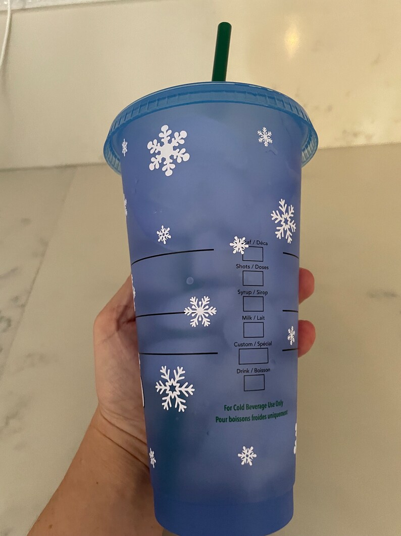 Puede incluir: Un vaso azul reutilizable para bebidas fr&iacute;as con dise&ntilde;os de copos de nieve blancos. El vaso tiene una pajita verde y una tapa azul. El texto en el vaso incluye "Shots/Doses", "Syrup/Sirop", "Milk/Lait", "Custom/Sp&eacute;cial" y "Drink/Boisson".