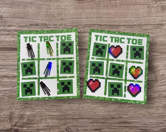 Op de afbeelding: Twee groene en witte Tic Tac Toe-spelborden met kleurrijke spelstukken. De spelstukken omvatten groene Creeper-gezichten, rode harten en blauwe en zwarte Minecraft-karakters.