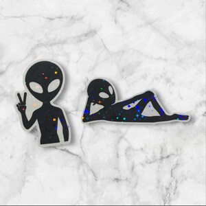 May include: Two black alien stickers with a glittery finish. One alien is standing with a peace sign and the other is lying down.