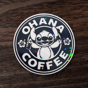May include: A round sticker with a black border and the words "OHANA COFFEE" in white. The sticker features a cartoon character holding a coffee cup. The character is white with large ears and a wide smile. The sticker has a holographic effect.