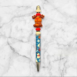 May include: A blue and white pen with a reindeer topper. The pen has a silver tip and a blue barrel with a white snowflake pattern. The reindeer is wearing a red hat and a brown scarf.