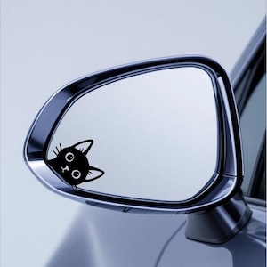 Puede incluir: Una pegatina de un gato negro en el espejo retrovisor lateral de un coche. Se ve la cabeza del gato, con ojos grandes y orejas puntiagudas. El espejo es cromado y refleja un cielo azul claro.