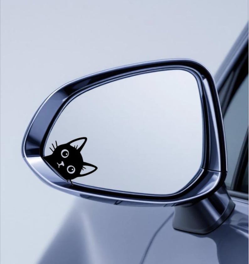 Puede incluir: Una pegatina de un gato negro en el espejo retrovisor lateral de un coche. Se ve la cabeza del gato, con ojos grandes y orejas puntiagudas. El espejo es cromado y refleja un cielo azul claro.