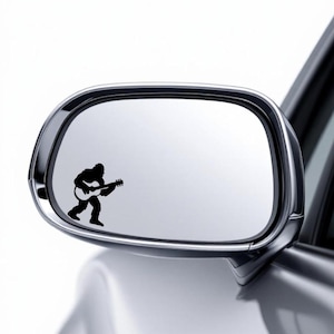 May include: A black vinyl decal of Bigfoot playing a guitar is affixed to a car's side mirror. The sticker is a silhouette of the legendary creature, holding the instrument. The mirror is chrome-trimmed and reflects the car's silver body.