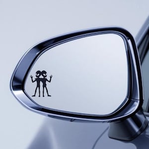 May include: A black vinyl decal of two cartoon aliens waving, applied to a car side mirror. The aliens have large eyes and slender bodies. The mirror is chrome and reflects a light blue sky.