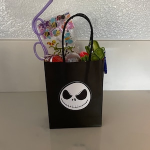 May include: A black paper gift bag with a white Jack Skellington face design. The bag is filled with various small toys and trinkets, including a purple bendy straw, a package of colorful stickers, a green fidget toy, and a red and white lollipop.