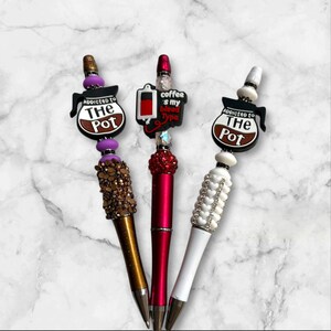 May include: Three decorative pens with coffee pot themed tops. The pens are gold, red, and white with rhinestones. The tops say "Addicted to the Pot" and "Coffee is my blood type".