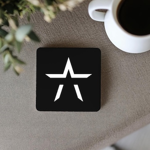 May include: A black square coaster with a white star design. The star has a central point and two downward-pointing lines. A white coffee mug filled with dark liquid is in the background.
