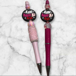 May include: Two decorative pens with the text "Mama Mommy Ma Bruh" on a black background. One pen is light pink with a crystal-studded top, and the other is dark pink. Both pens have silver tips and decorative beads.