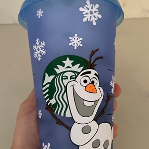 Puede incluir: Un vaso azul de Starbucks con copos de nieve blancos y una imagen de dibujos animados de Olaf de Frozen. El vaso tiene una pajita verde y el logotipo de Starbucks. La taza est&aacute; sujeta por una mano.