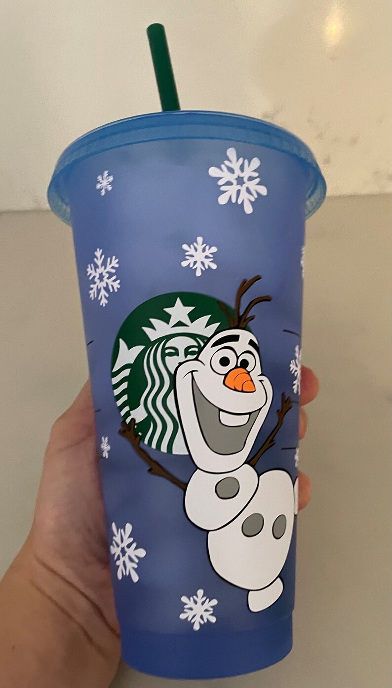 Puede incluir: Un vaso azul de Starbucks con copos de nieve blancos y una imagen de dibujos animados de Olaf de Frozen. El vaso tiene una pajita verde y el logotipo de Starbucks. La taza est&aacute; sujeta por una mano.