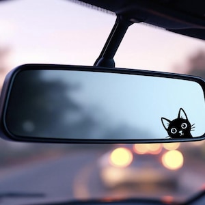 May include: A black cat decal is affixed to a car's rearview mirror. The cat's head is peeking out, with large eyes and a curious expression. The background shows a blurred view of the road.