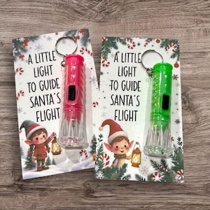 May include: Two Christmas-themed keychains. One is pink, the other green, each with a small light and the text "A LITTLE LIGHT TO GUIDE SANTA'S FLIGHT." The cards feature an elf illustration and festive decorations.