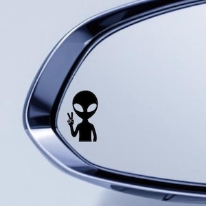 May include: Black vinyl decal of an alien giving the peace sign. The alien has a large head, oval eyes, and a small body. The decal is shown on a car side mirror.