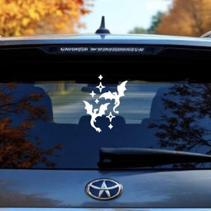 May include: White vinyl car decal featuring a cluster of stylized bats and stars. The decal is applied to the rear window of a gray vehicle. The background includes fall foliage.