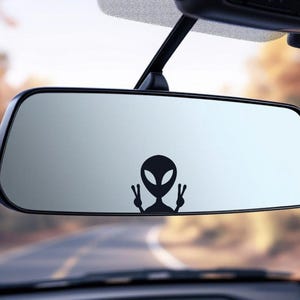 May include: A black alien decal with a peace sign gesture is affixed to a car's rearview mirror. The mirror reflects a blurred outdoor scene with a road and trees. The alien has large oval eyes and a small head.