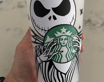 May include: A white reusable cup with a black and white illustration of Jack Skellington from The Nightmare Before Christmas holding a green Starbucks logo. The cup has a black lid.