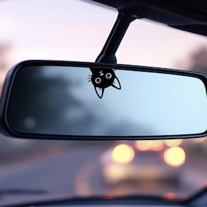 Puede incluir: Una calcoman&iacute;a de gato negro est&aacute; adherida al espejo retrovisor de un coche. La cabeza del gato, con grandes ojos y orejas puntiagudas, es visible. El espejo refleja una vista borrosa de la carretera y otros veh&iacute;culos.