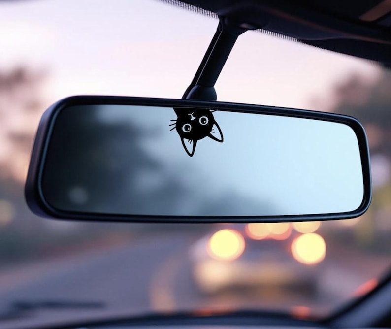 Puede incluir: Una calcoman&iacute;a de gato negro est&aacute; adherida al espejo retrovisor de un coche. La cabeza del gato, con grandes ojos y orejas puntiagudas, es visible. El espejo refleja una vista borrosa de la carretera y otros veh&iacute;culos.