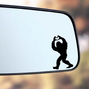 May include: A black silhouette of Bigfoot, also known as Sasquatch, is displayed on a car side mirror. The figure is in a walking pose, with arms raised. The mirror has a reflective surface and a black frame.