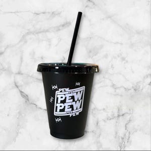 May include: Black reusable cold cup with a black lid and straw. The cup features the text "PEW PEW" in a white rectangular design, along with white illustrations of a starfighter. A fun, reusable cup for cold beverages.