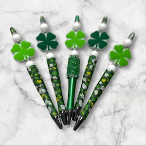 May include: A collection of decorative pens with green and white floral patterns. Each pen has a silver tip and a clover-shaped charm in shades of green. One pen is covered in green glitter.