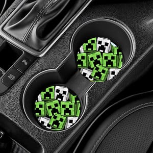 May include: Two car coasters with a green and black pixelated design of a Minecraft Creeper face. The coasters are round and sit inside the car's cup holders. The car's interior is black.