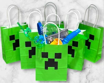 Op de afbeelding: Vijf groene papieren cadeauzakjes met witte handvatten, elk met een zwart gepixeld gezichtsontwerp. De tassen zijn gevuld met diverse speelgoed en accessoires, waaronder een blauwe armband, een speelgoedauto en een blauw waterpistool.