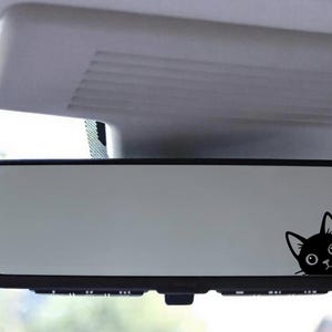 Puede incluir: Una pegatina de un gato negro asom&aacute;ndose por el espejo retrovisor de un coche. El gato tiene un dise&ntilde;o de dibujos animados con ojos grandes y una expresi&oacute;n de sorpresa. El espejo es rectangular con un marco negro.