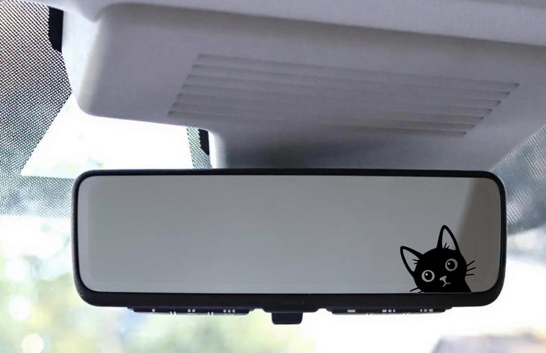 Puede incluir: Una pegatina de un gato negro asom&aacute;ndose por el espejo retrovisor de un coche. El gato tiene un dise&ntilde;o de dibujos animados con ojos grandes y una expresi&oacute;n de sorpresa. El espejo es rectangular con un marco negro.