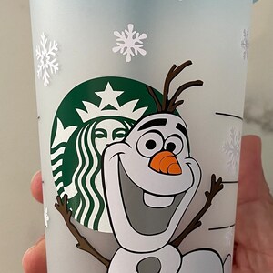 Puede incluir: Un vaso fr&iacute;o de Starbucks de pl&aacute;stico transparente con tapa azul claro y pajita verde. La taza presenta una imagen de dibujos animados de Olaf el mu&ntilde;eco de nieve de Frozen, junto con calcoman&iacute;as de copos de nieve blancos. El logotipo de Starbucks tambi&eacute;n es visible.