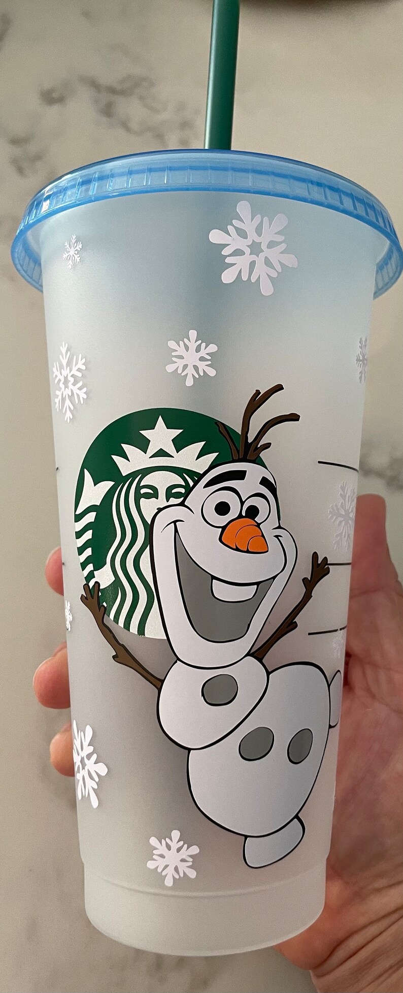 Puede incluir: Un vaso fr&iacute;o de Starbucks de pl&aacute;stico transparente con tapa azul claro y pajita verde. La taza presenta una imagen de dibujos animados de Olaf el mu&ntilde;eco de nieve de Frozen, junto con calcoman&iacute;as de copos de nieve blancos. El logotipo de Starbucks tambi&eacute;n es visible.