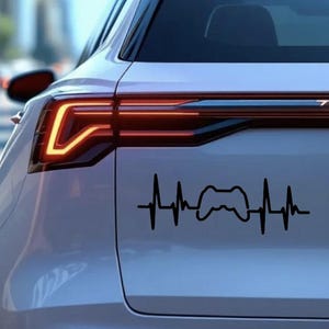 May include: A black vinyl decal of a video game controller integrated into a heartbeat design, applied to the rear of a white car. The car's taillight is visible. The design suggests a gaming theme.