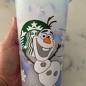 Puede incluir: Un vaso fr&iacute;o de Starbucks de pl&aacute;stico transparente con tapa y pajita azules. La taza presenta a Olaf de Frozen sonriendo, copos de nieve y el logotipo de Starbucks. La taza est&aacute; dise&ntilde;ada para bebidas fr&iacute;as.