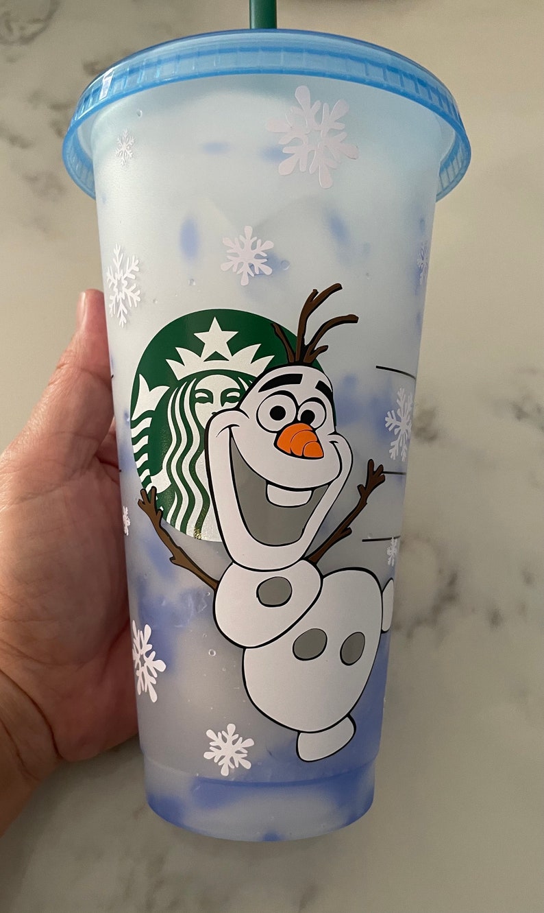 Puede incluir: Un vaso fr&iacute;o de Starbucks de pl&aacute;stico transparente con tapa y pajita azules. La taza presenta a Olaf de Frozen sonriendo, copos de nieve y el logotipo de Starbucks. La taza est&aacute; dise&ntilde;ada para bebidas fr&iacute;as.