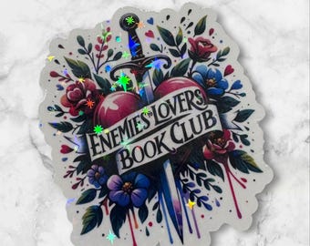 May include: A white sticker with a colorful illustration of a heart pierced by a sword. The heart is surrounded by flowers and the text "Enemies to Lovers Book Club" is written on a banner.