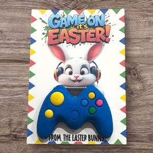 May include: A blue and yellow game controller with a white bunny wearing headphones on top. The card says "Game On It's Easter! From, The Easter Bunny".