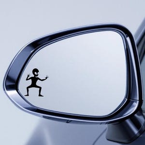 May include: A black vinyl decal of an alien figure is affixed to a car side mirror. The alien has a large head, small body, and outstretched arms and legs. The mirror is chrome and reflects a light blue background.
