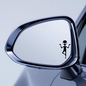 May include: A black alien decal is affixed to a car's side mirror. The decal depicts a stylized alien figure with arms raised, standing on one leg. The mirror has a chrome frame and reflects a light blue sky.