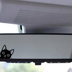 Puede incluir: Una calcoman&iacute;a de gato negro est&aacute; adherida al espejo retrovisor de un autom&oacute;vil. La cabeza del gato es visible, con ojos grandes y redondos y orejas puntiagudas. La calcoman&iacute;a agrega un toque l&uacute;dico al interior del veh&iacute;culo.