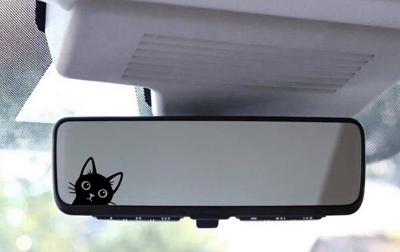 Puede incluir: Una calcoman&iacute;a de gato negro est&aacute; adherida al espejo retrovisor de un autom&oacute;vil. La cabeza del gato es visible, con ojos grandes y redondos y orejas puntiagudas. La calcoman&iacute;a agrega un toque l&uacute;dico al interior del veh&iacute;culo.