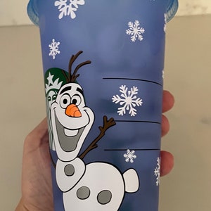 Puede incluir: Un vaso de pl&aacute;stico azul con una tapa a juego y una pajita verde. El vaso presenta una imagen de dibujos animados de Olaf de Frozen, junto con dise&ntilde;os de copos de nieve blancos. El logotipo de Starbucks es visible.