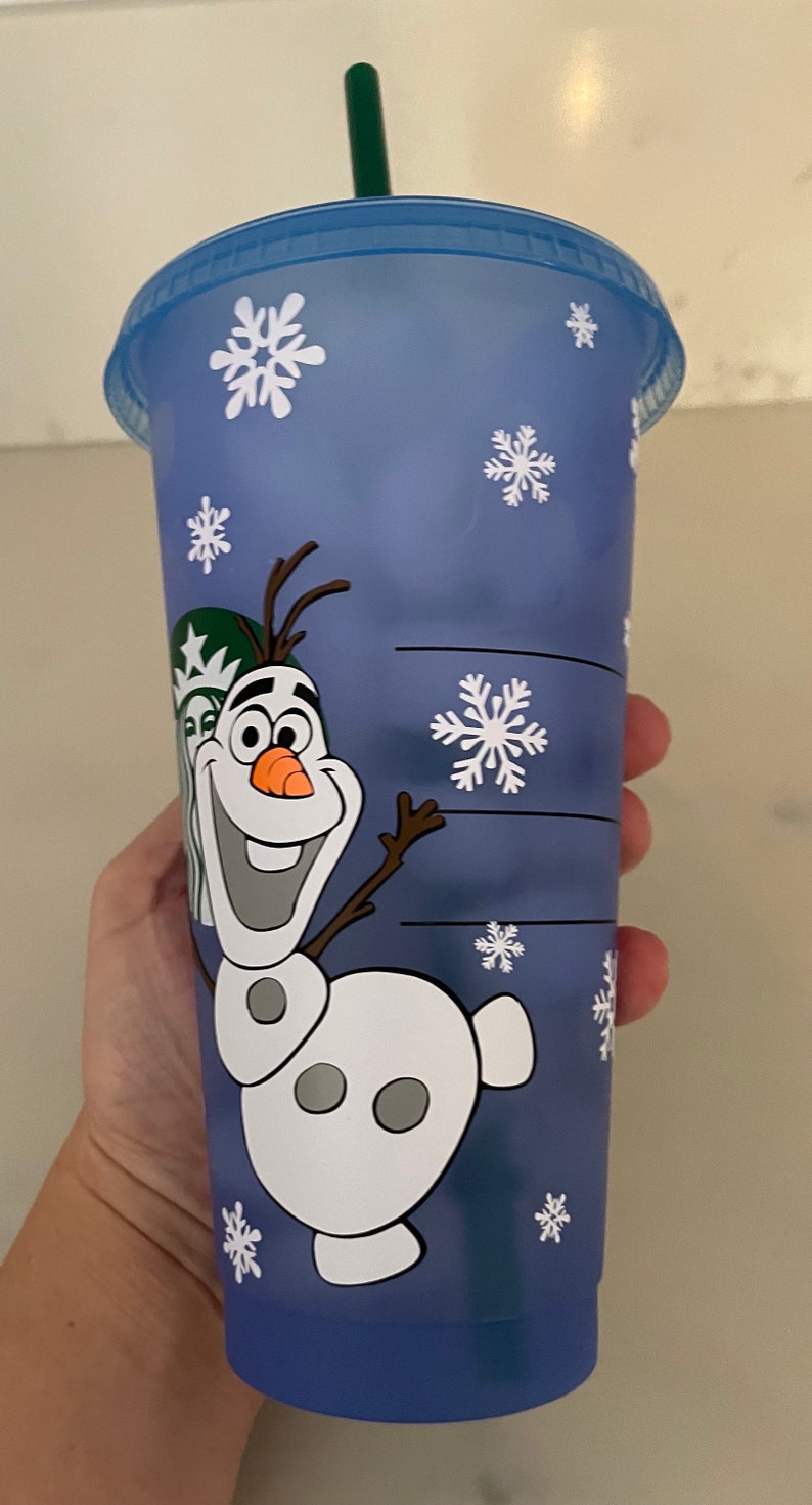 Puede incluir: Un vaso de pl&aacute;stico azul con una tapa a juego y una pajita verde. El vaso presenta una imagen de dibujos animados de Olaf de Frozen, junto con dise&ntilde;os de copos de nieve blancos. El logotipo de Starbucks es visible.
