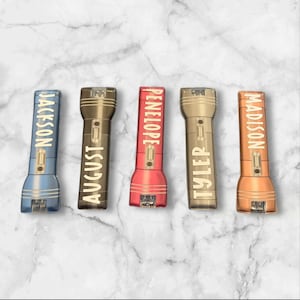 May include: Five personalized flashlights in blue, bronze, red, gold, and copper. Each flashlight has a name printed vertically in gold: Jackson, August, Penelope, Tyler, and Madison. The flashlights are arranged in a row against a marble background.