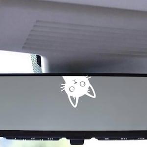 Puede incluir: Retrovisor de coche con una pegatina de cara de gato blanco. La cabeza del gato est&aacute; boca abajo, con ojos grandes y orejas puntiagudas. El espejo es rectangular con un marco negro, y el fondo muestra el interior del coche.
