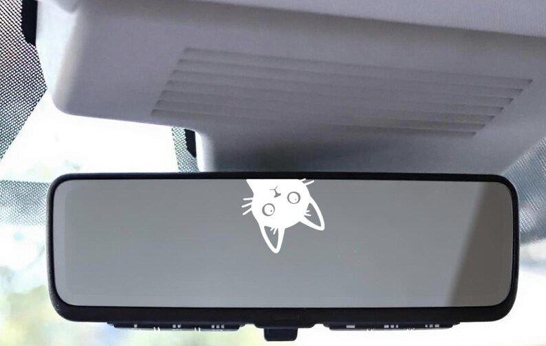 Puede incluir: Retrovisor de coche con una pegatina de cara de gato blanco. La cabeza del gato est&aacute; boca abajo, con ojos grandes y orejas puntiagudas. El espejo es rectangular con un marco negro, y el fondo muestra el interior del coche.
