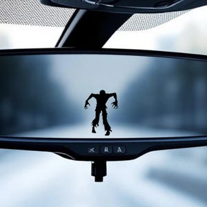 May include: Black silhouette of a zombie figure, with outstretched arms and tattered clothing, is affixed to a car's rearview mirror. The mirror reflects a blurred, overcast outdoor scene, creating a spooky, Halloween-themed aesthetic.
