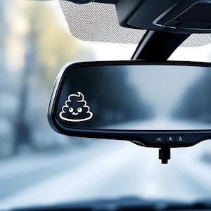 May include: A white, cartoon-style poop emoji sticker is affixed to a car's rearview mirror. The emoji has a smiling face and is outlined in white. The mirror is black, and the background is blurred.
