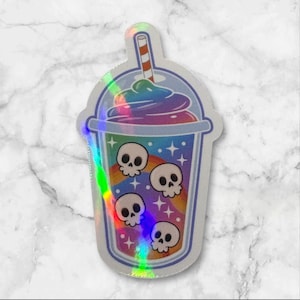 May include: A holographic sticker in the shape of a beverage cup. The cup is filled with a rainbow-colored drink and topped with a swirl of whipped cream. Four white skulls are on a rainbow background. A red and white striped straw is in the drink.